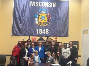 A group of people standing in front of a large Wisconsin state flag in the U.S. Capitol.