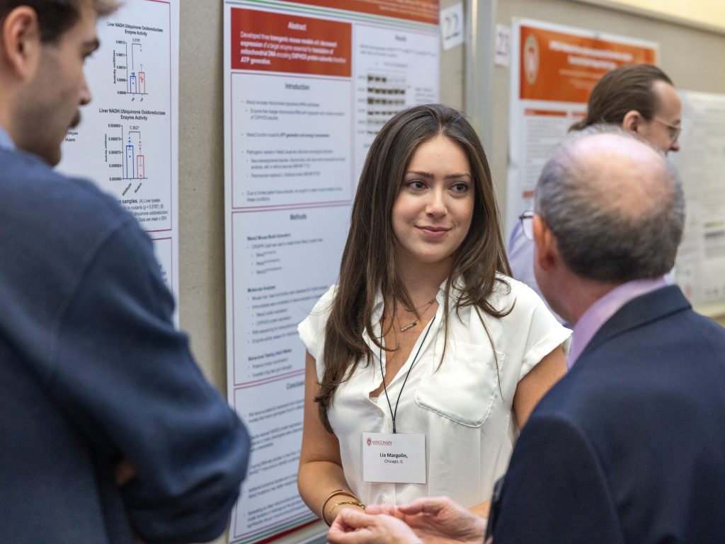 Lia Margolin standing in front of her poster in discussion with two men with their backs to the camera at the Precision Medicine Symposium 2025
