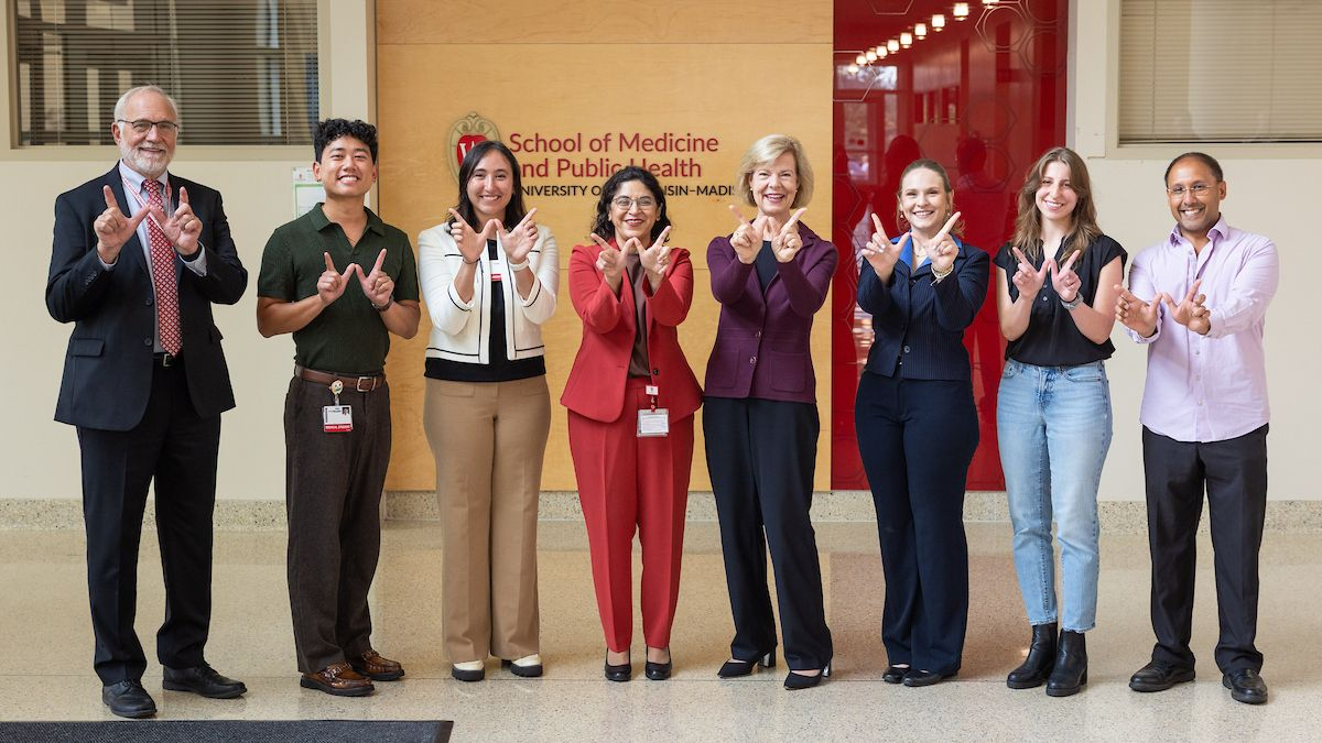 Senator Tammy Baldwin; UW-Madison School of Medicine and Public Health Dean Dr. Nita Ahuja, MD, MBA; and several UW-Madison students standing and smiling together while making "Ws" with their hands