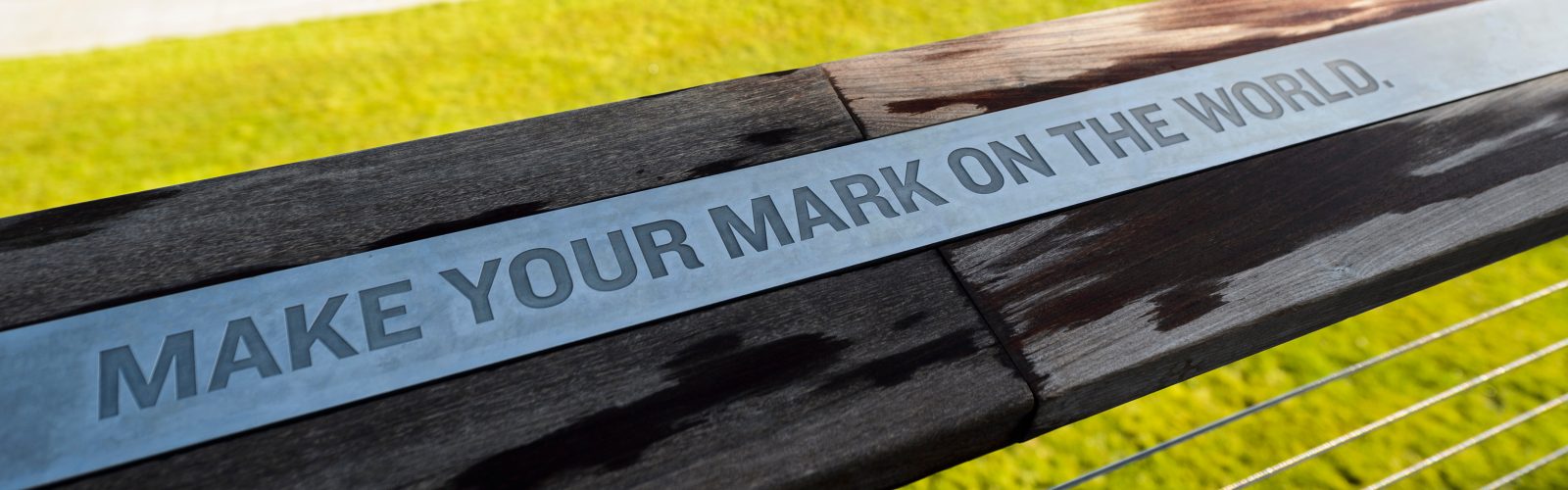 Metal plaque on a wooden railing with the engraved words “MAKE YOUR MARK ON THE WORLD.” against a blurred grassy background.
