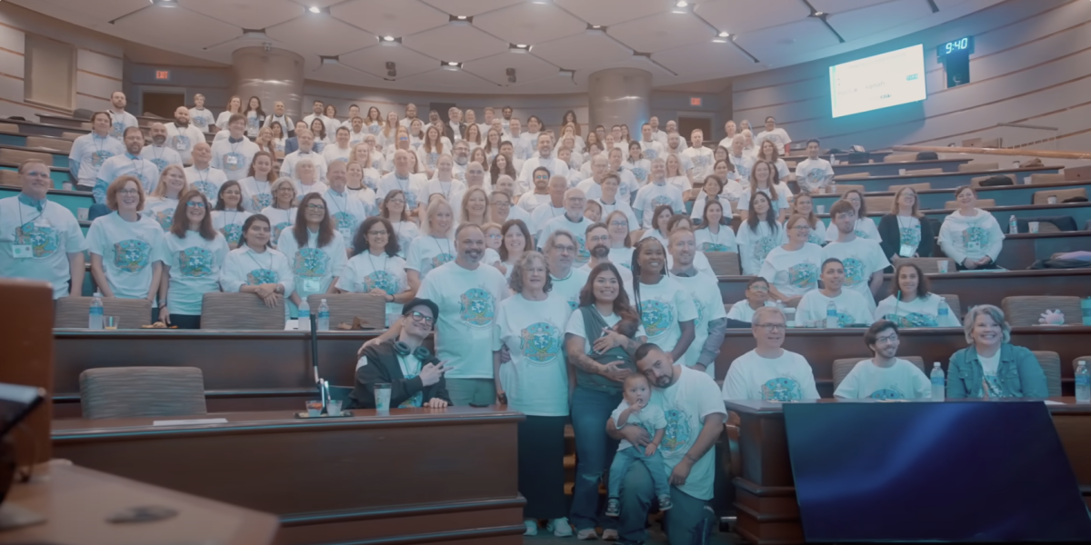A lecture hall full of smiling people wearing the same white t-shirt with a logo on it posing for a photo for the Undiagnosed Hackathon 2025