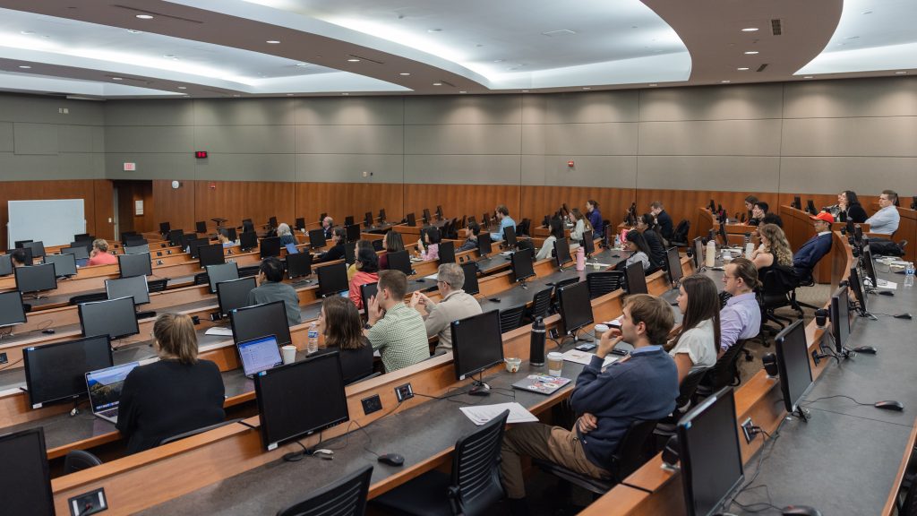 a large lecture hall with built in computer monitors mostly filled with people in attendance at the Precision Medicine Symposium 2025
